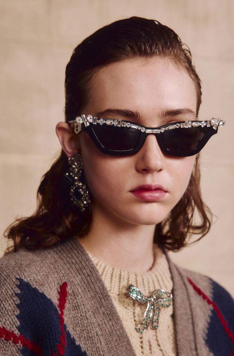 Woman wearing sunglasses adorned with rhinestones, a light beige sweater, and statement earrings, posing against a neutral background.