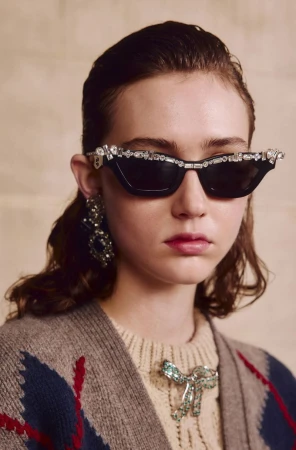 Woman wearing sunglasses adorned with rhinestones, a light beige sweater, and statement earrings, posing against a neutral background.