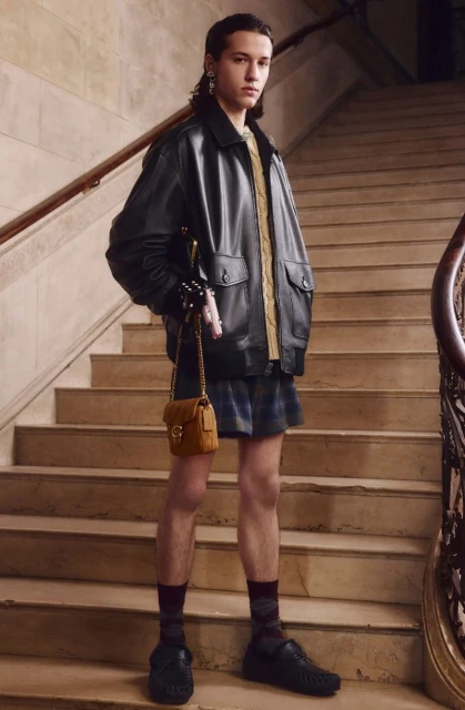 Male fashion model poses on staircase wearing a black leather jacket, plaid shorts, gold sweater and carrying a brown cross-body bag.