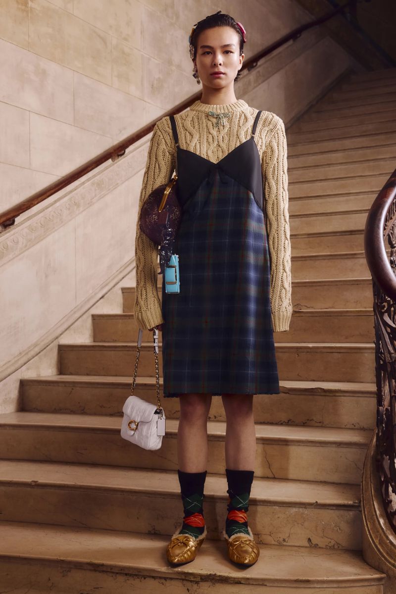 Full-length studio shot of a woman wearing a slip dress and cream-colored sweater, accessorized with two handbags, on a staircase.