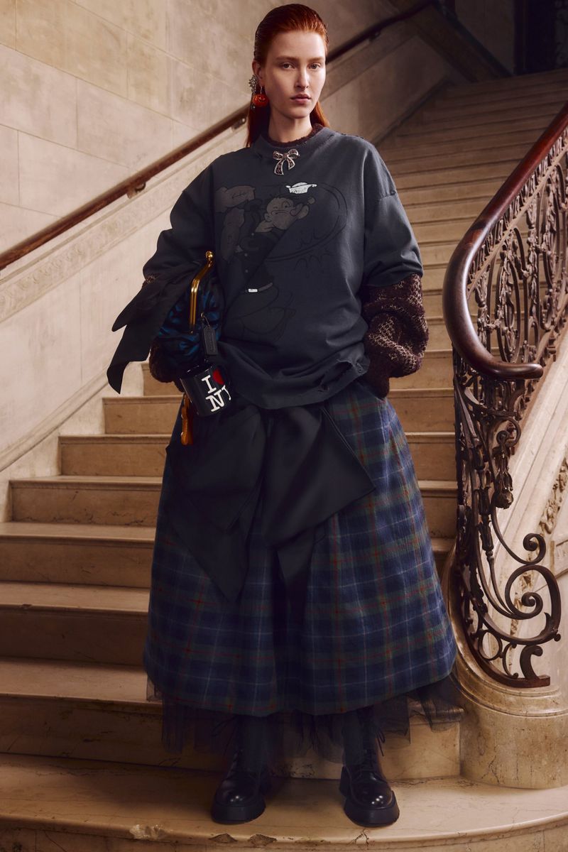 Fashionable redhead woman on marble stairs in a grey graphic sweatshirt, plaid skirt, accessorized with statement earrings and handbag.