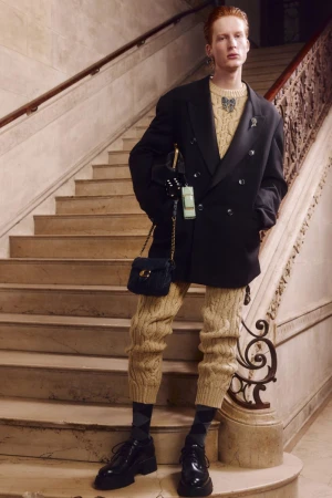 Man in a black double-breasted blazer, light-brown cable-knit sweater, brown cable-knit pants, socks, and chunky shoes, posing on marble steps.