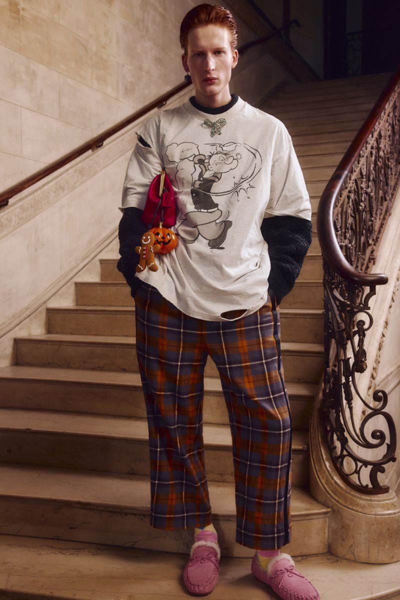 Male fashion model poses on marble staircase wearing graphic t-shirt, plaid pants, and moccasin slippers. Indoor shot, natural light.
