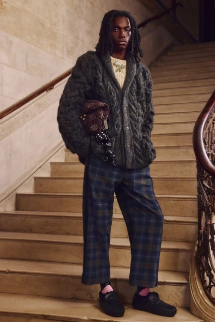 Young adult stands on light-brown stairs with patterned bag and clothing. The person wears a gray cardigan and blue plaid pants.