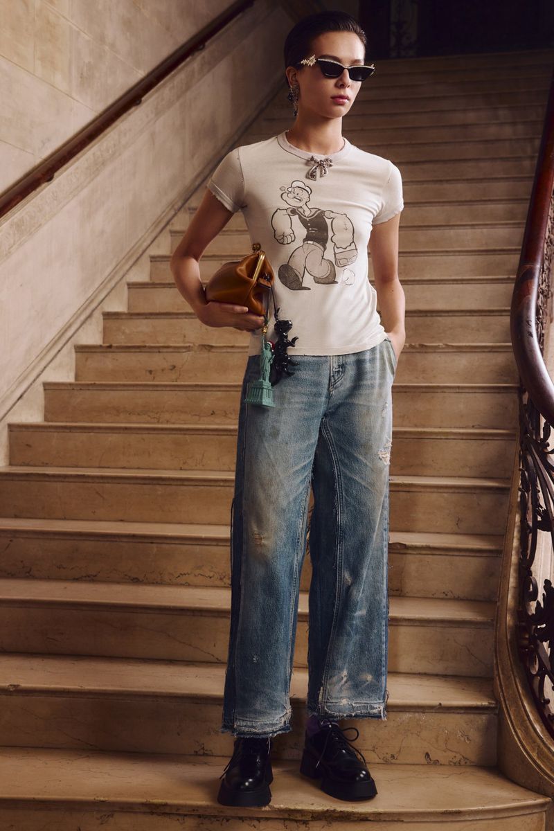 Model wearing a graphic tee, cat-eye sunglasses, and wide-leg denim jeans on a stone staircase for Coach.