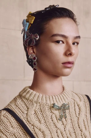 Close-up of a model with jeweled hair clips and earring wearing a tan cable knit sweater posing indoors against a neutral backdrop.