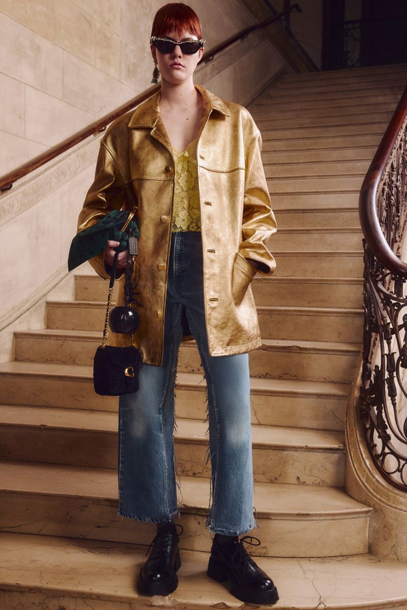 Woman in golden coat and jeans stands on stairs holding purse and pouch. Fashionable street style portrait with sunglasses and black boots.