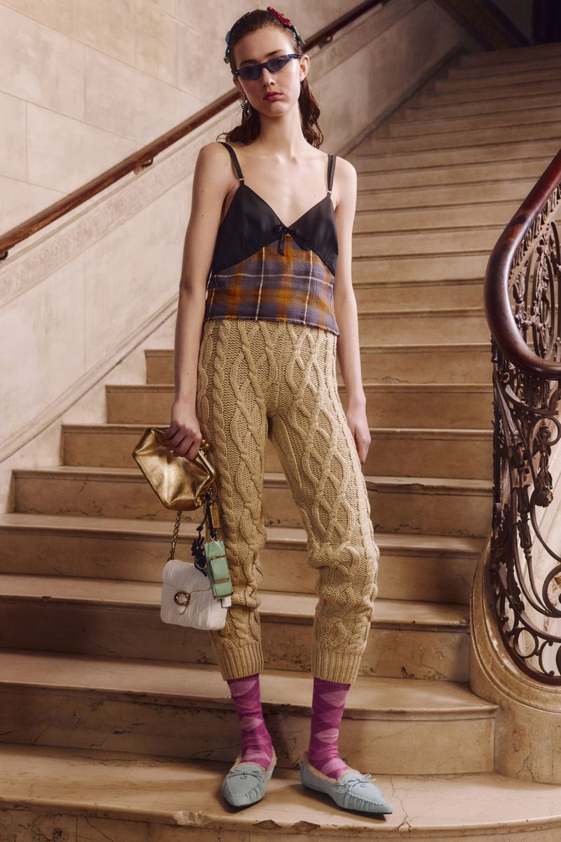 Indoor shot of a woman in a black and plaid top and knit pants posing on stairs with handbags. Fashion editorial lifestyle photography.