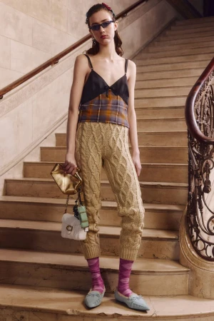 Indoor shot of a woman in a black and plaid top and knit pants posing on stairs with handbags. Fashion editorial lifestyle photography.