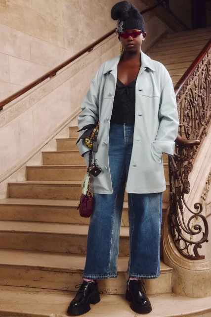 Stylish woman poses on staircase wearing a blue trench coat, wide leg jeans, and chunky platform shoes with handbag accessory.