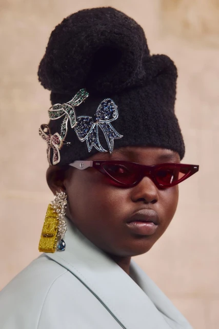 Woman with dark skin and red glasses wearing a black knit hat adorned with jewel-toned bows and a statement earring, fashion shot.
