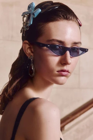 Close-up of an adult female fashion model with sunglasses, blue hair accessories, and teardrop earrings looking back over her shoulder.