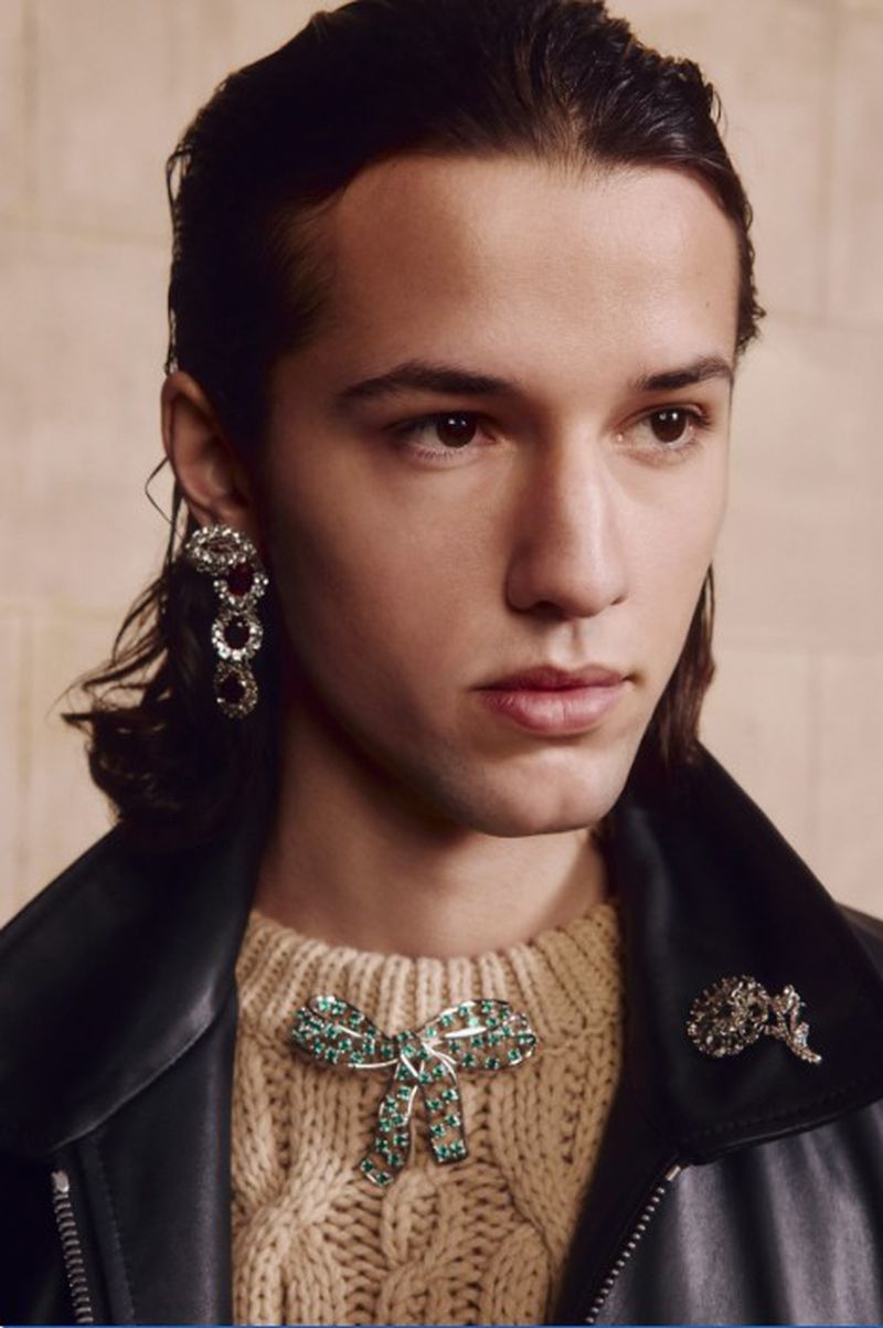 Close-up studio shot of a young adult man wearing a tan cable knit sweater, black leather jacket, and green jewel-encrusted brooches and earrings.