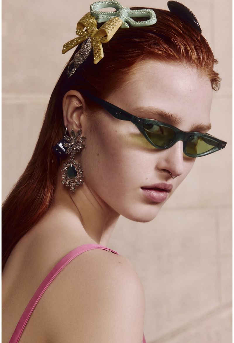 Close-up shot of a red-haired woman wearing retro green sunglasses, large ornamental earrings, and ribbon-shaped barrettes.