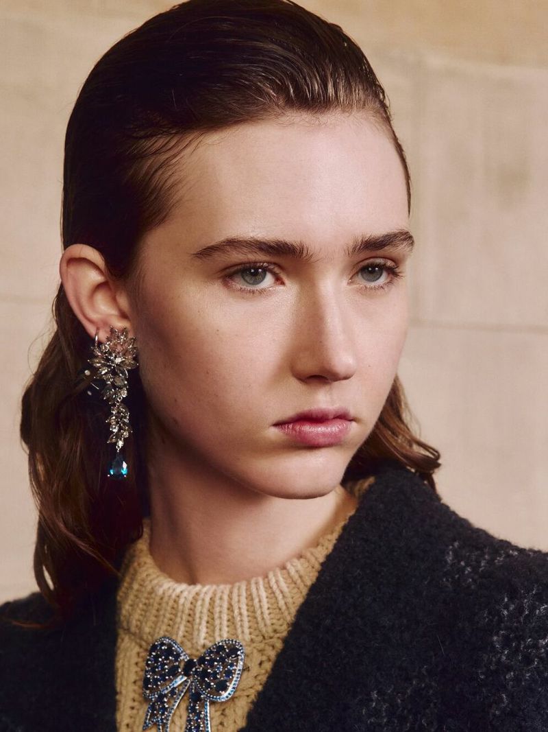 Close-up shot of a woman wearing ornate jewel earrings and a bow brooch, featuring light skin, brown hair, and light makeup. 