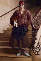 Model standing on a grand staircase wearing a chunky brown cable-knit cardigan and dark sunglasses, holding a brown leather bag