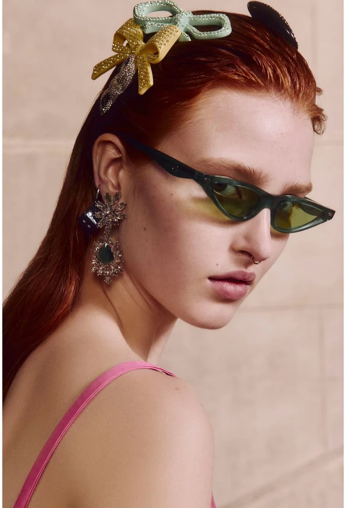 Close-up portrait of a red-haired woman wearing green cat-eye sunglasses, crystal earrings, and colorful bow hair clips for Coach Resort 2025.