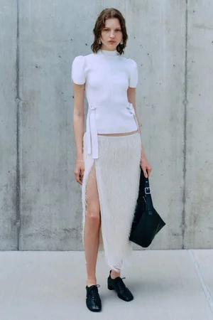 Model wearing white top and fringed skirt standing against concrete wall for 3.1 Phillip Lim Pre-Spring 2025.
