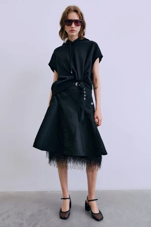 Model wearing a black hooded top and flared skirt with sunglasses against a seamless white studio background