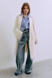 Model wearing a fringed white coat, striped button-down shirt, and wide-leg denim jeans against a plain white background.