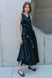 Model Nadine Kirilova in a sleeveless black satin asymmetrical dress and mesh block heels standing against a curved concrete wall.