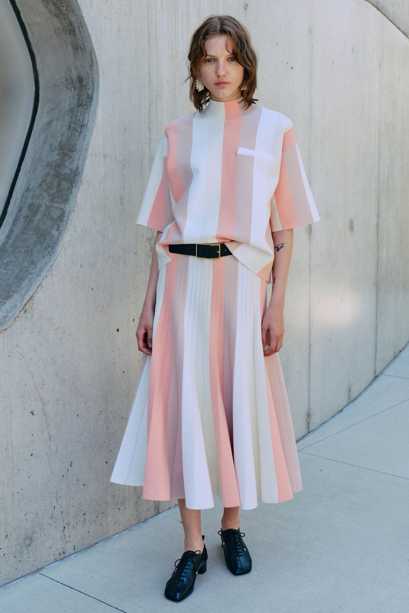 Woman poses in a peach and white striped top and skirt with a black belt, set against a textured grey wall in natural light.