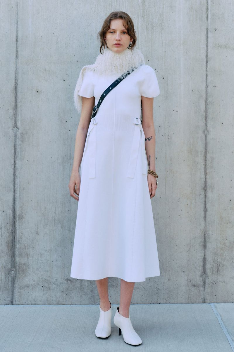 A young adult female stands in a white dress and booties against a concrete background, wearing a furry collar and carrying a bag.