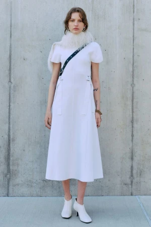 A young adult female stands in a white dress and booties against a concrete background, wearing a furry collar and carrying a bag.