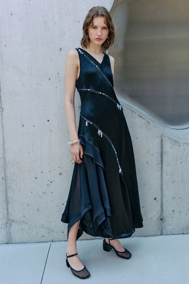 An adult woman poses in an elegant black sleeveless dress with embellishments, complemented by dark shoes, against a concrete background.