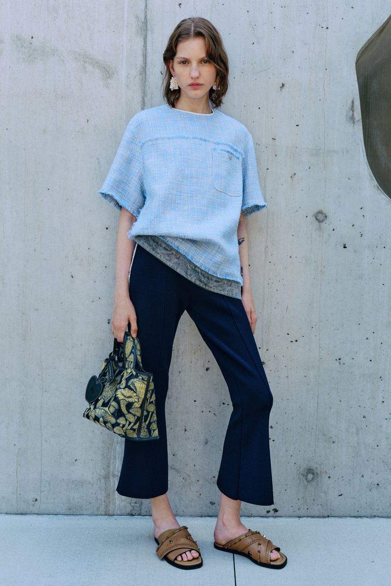 A woman in a light-blue tweed top, dark trousers, and sandals poses against a concrete wall with a patterned handbag, showcasing a fashion-forward look.
