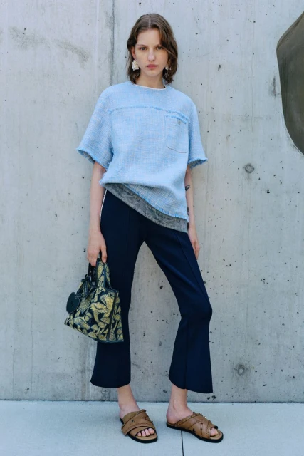 A woman in a light-blue tweed top, dark trousers, and sandals poses against a concrete wall with a patterned handbag, showcasing a fashion-forward look.