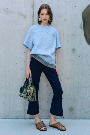 A woman in a light-blue tweed top, dark trousers, and sandals poses against a concrete wall with a patterned handbag, showcasing a fashion-forward look.