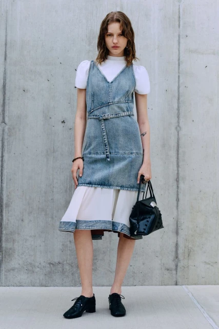 A fair-skinned young adult woman stands against a concrete wall modeling a denim dress with white underskirt and white top with puff sleeves.