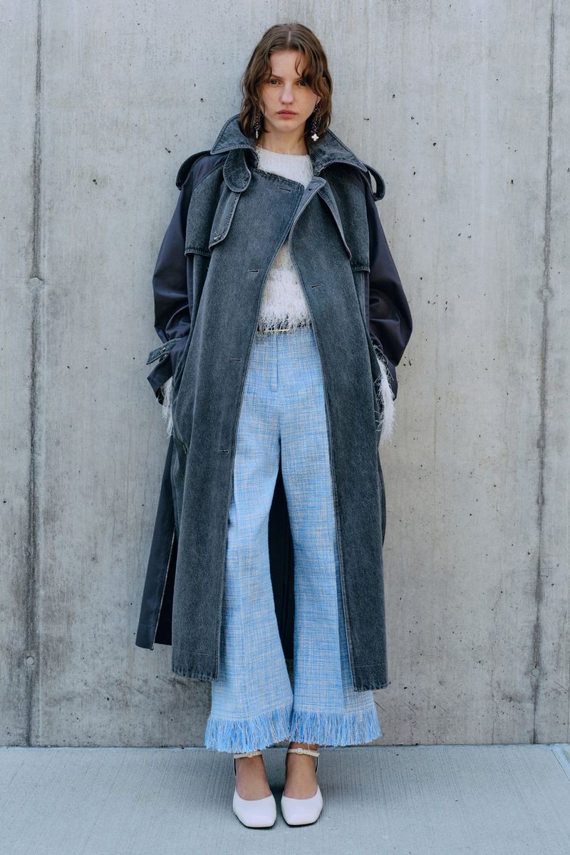 Adult female fashion model poses in a grey trench coat and light blue tweed pants against a concrete wall outdoors in daylight.