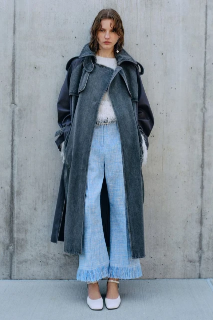 Adult female fashion model poses in a grey trench coat and light blue tweed pants against a concrete wall outdoors in daylight.