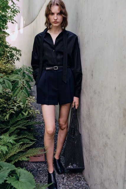 A slender woman stands against a concrete wall wearing a dark button-down shirt, shorts, and loafers, holding a black net bag.