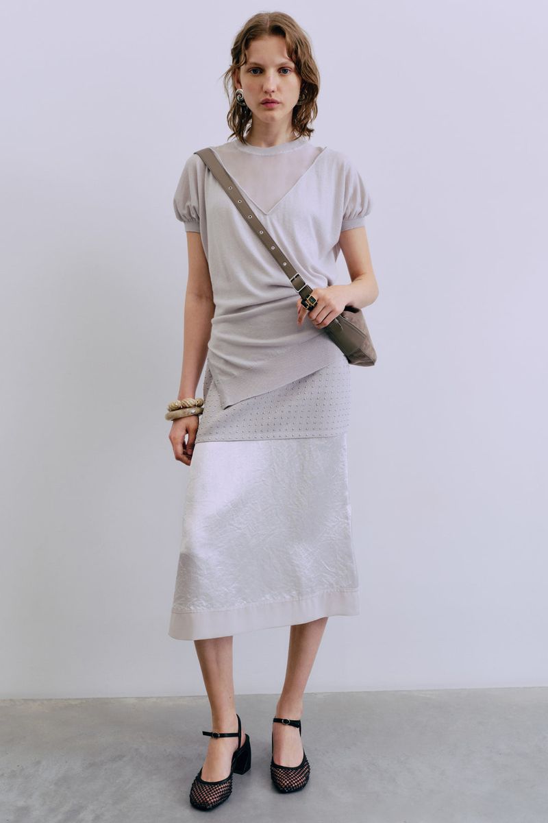 A woman poses in a fashionable light gray top and patterned skirt, accessorized with a shoulder bag and neutral-toned jewelry in studio lighting.