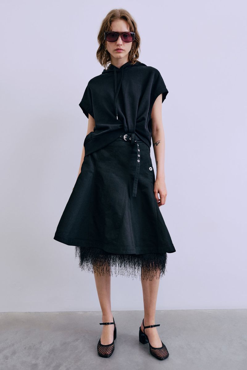Woman in a black hooded top and wrap skirt with fringe detail standing in front of a white background in a studio setting.