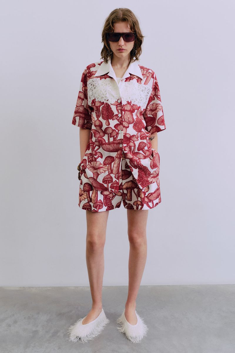 Young woman poses in mushroom-patterned shirt and shorts with white lace details and white furry shoes in a minimalist studio setting.