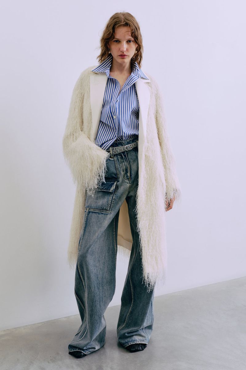 A fair-skinned young woman poses in a long white faux fur coat, blue striped shirt, and faded jeans against a plain white background.