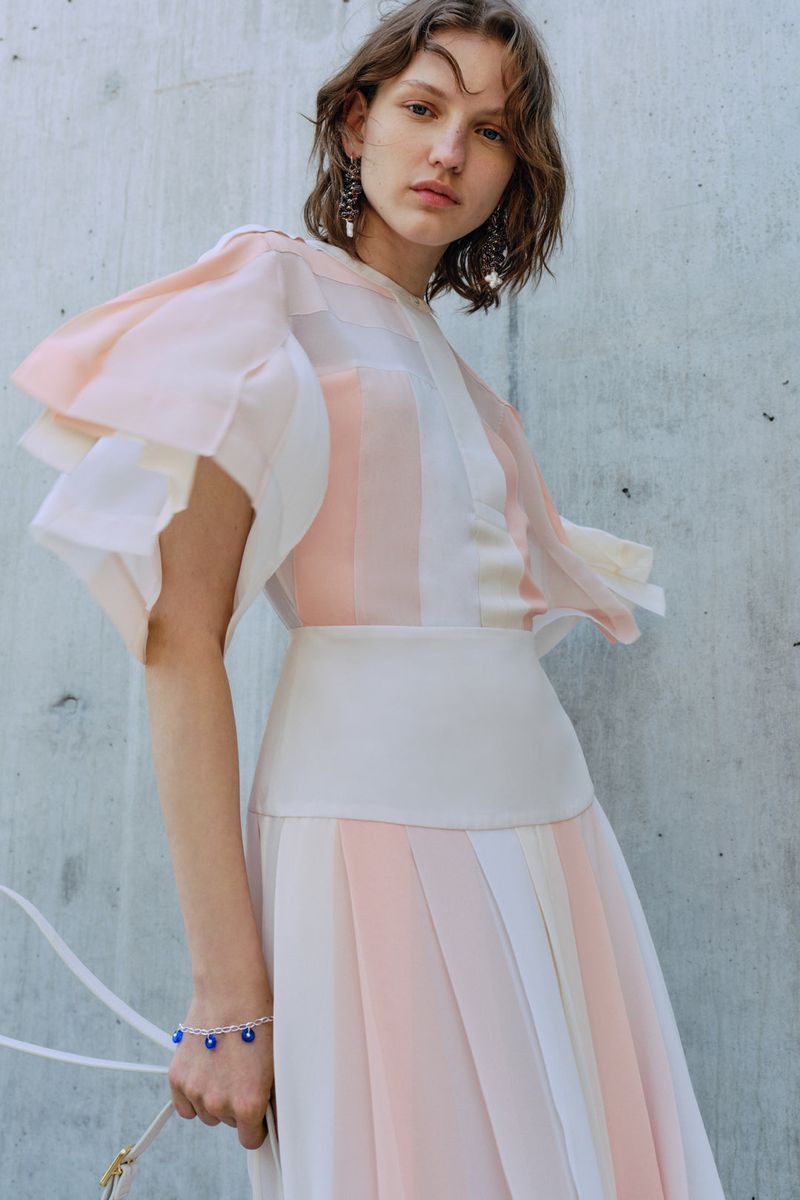 Woman in a flowing, layered dress with pastel stripes poses against a concrete wall, showcasing fashion and style in an outdoor setting.