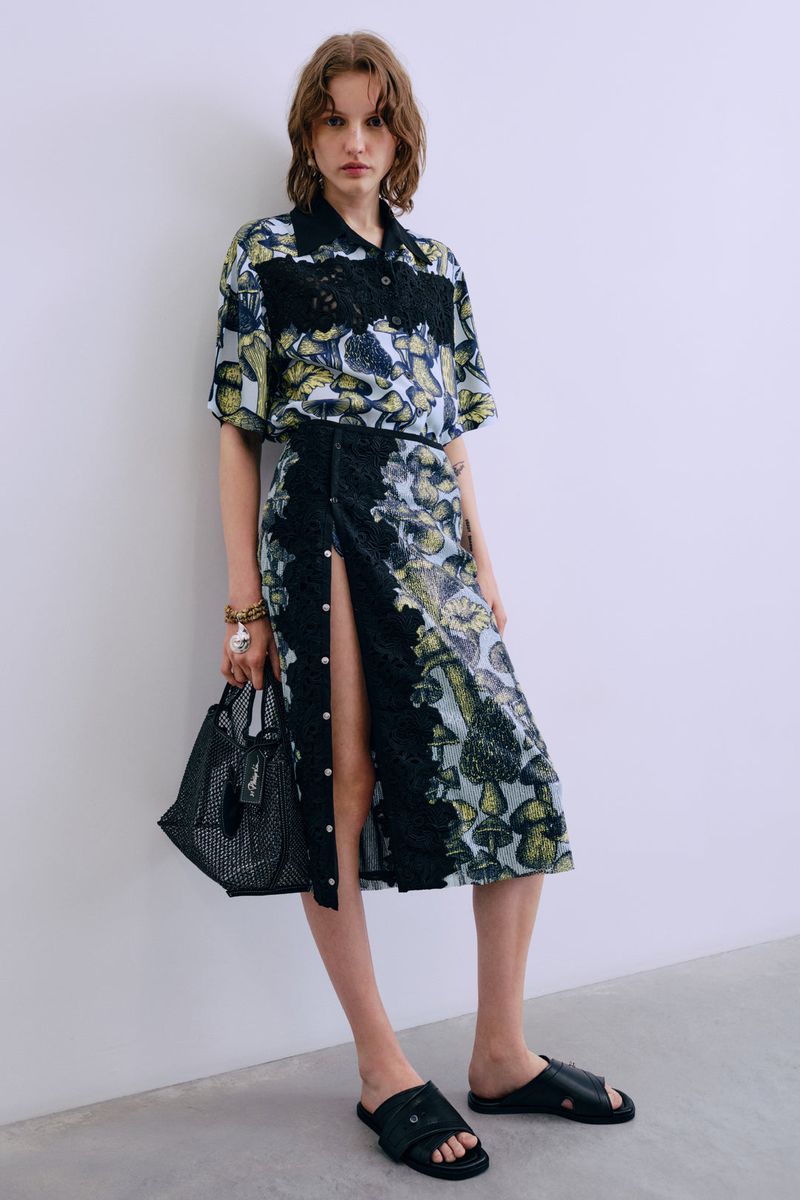 Fashion model wears a floral shirt and skirt ensemble in a studio setting, complemented by a black purse and stylish black sandals.