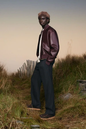 Full-length shot of a Black male model in a burgundy bomber jacket, cap, and dark pants, posing outdoors on grassy terrain.