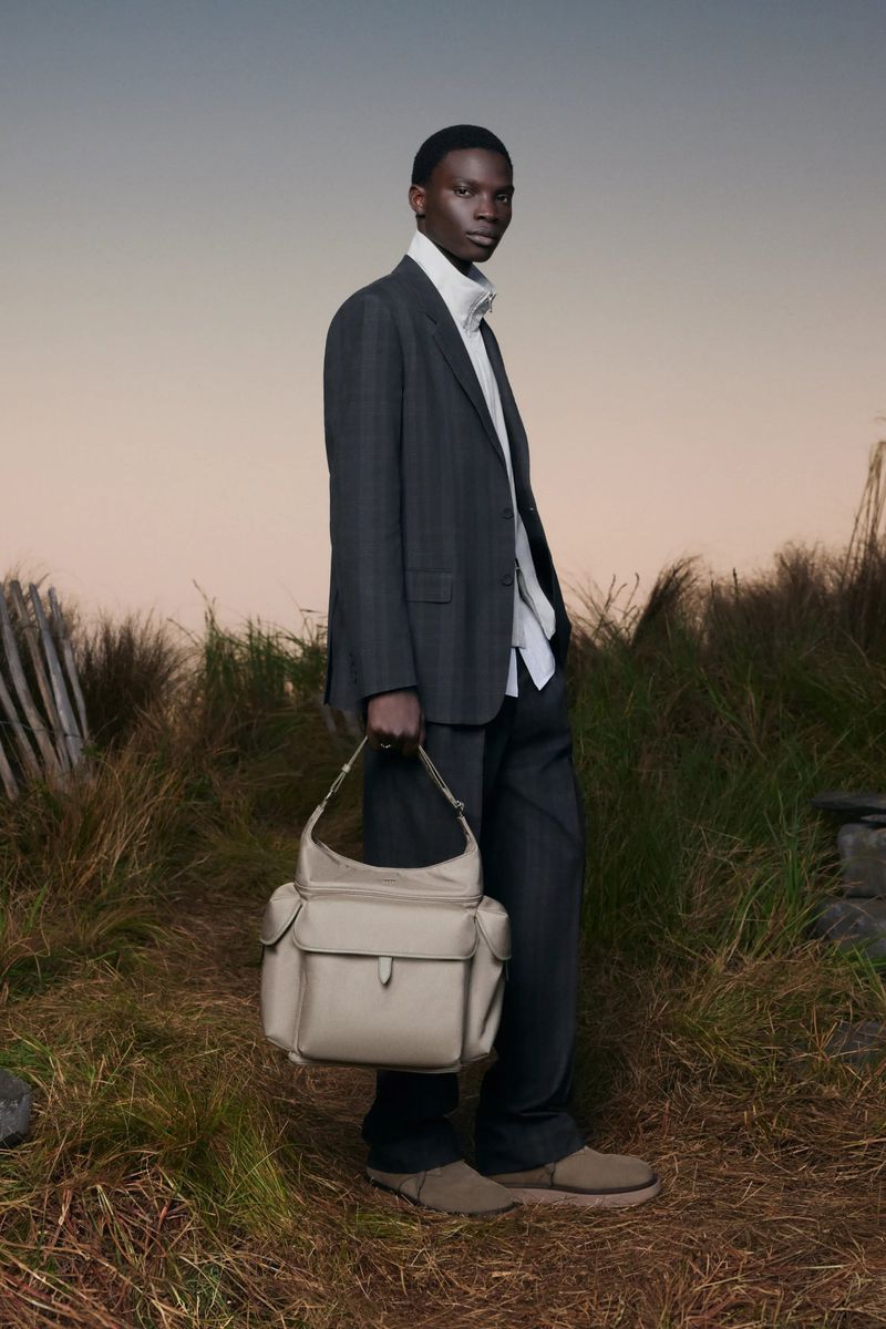 Fashionable adult Black male poses in a grey suit, cream turtleneck, holding a cream-colored handbag against an outdoor background.