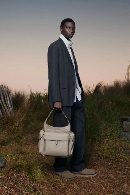 Fashionable adult Black male poses in a grey suit, cream turtleneck, holding a cream-colored handbag against an outdoor background.