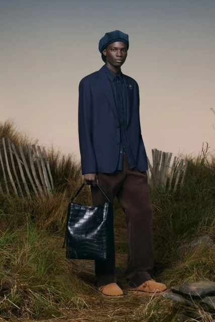 Dark-skinned male model wears navy blazer and beret, carrying black tote bag in a grassy field with light sky.