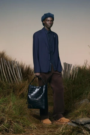 Dark-skinned male model wears navy blazer and beret, carrying black tote bag in a grassy field with light sky.