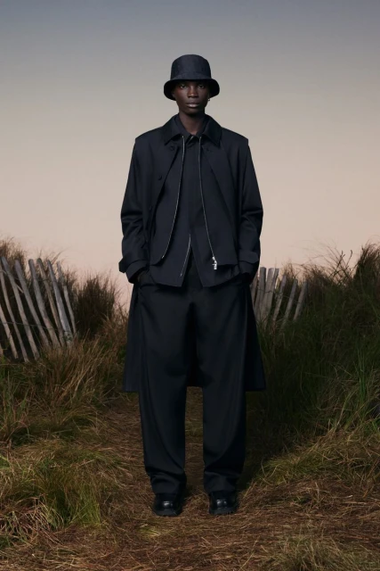 Man in dark, fashionable outerwear and bucket hat stands in tall grass beside rustic fence under a muted sky. Fashion editorial.