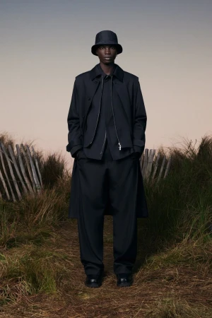 Man in dark, fashionable outerwear and bucket hat stands in tall grass beside rustic fence under a muted sky. Fashion editorial.