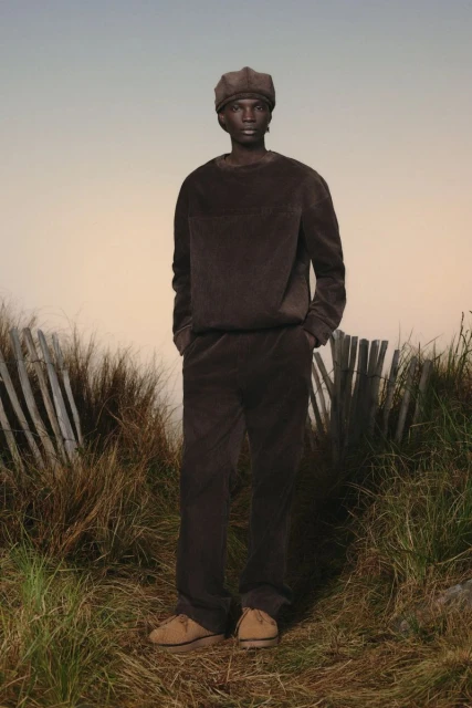 A Black male fashion model stands outdoors on a grassy hill wearing a brown corduroy outfit with a matching hat and suede shoes.